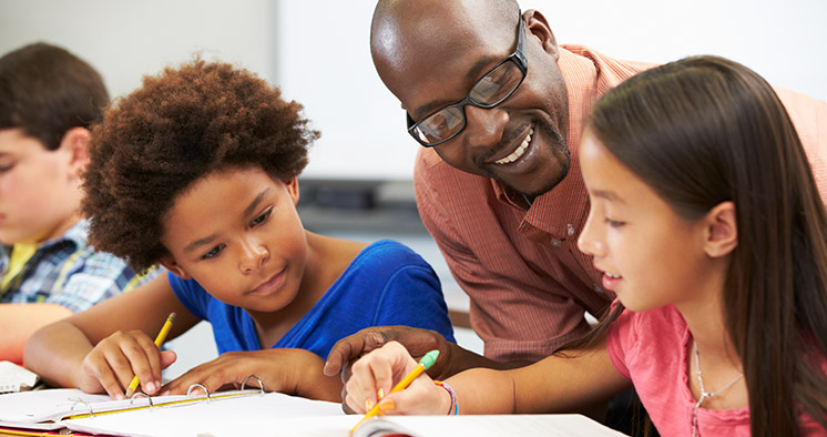 Teacher Helping Pupils Studying At Desks In Classroom